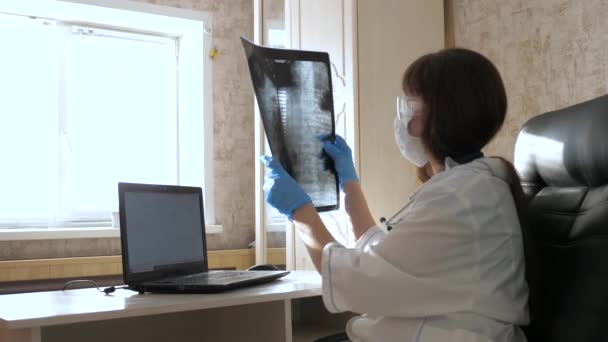 Μια γυναίκα γιατρός σε ένα γραφείο εξετάζει μια ακτινογραφία του ασθενούς στο νοσοκομείο. Ο γιατρός δουλεύει στο νοσοκομείο με λάπτοπ. Ιατρική περίθαλψη και υγειονομική περίθαλψη έννοια. πανδημία του κορονοϊού, πνευμονία COVID-19 — Αρχείο Βίντεο
