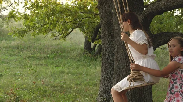 Anak-anak berayun di dahan pohon oak di bawah sinar matahari. Mimpi untuk terbang. konsep masa kecil yang bahagia. Gadis-gadis cantik bermain di taman. gadis remaja menikmati terbang pada ayunan pada malam musim panas di hutan — Stok Foto