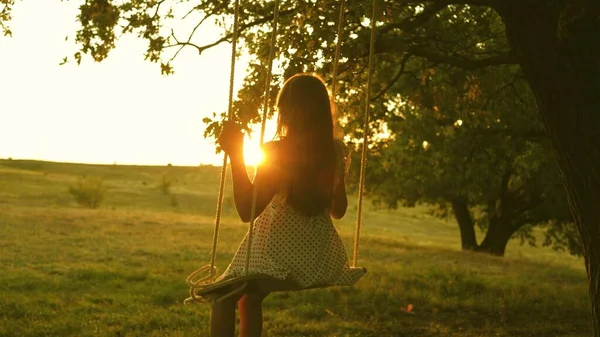 Bambino che oscilla su un'altalena nel parco al sole. giovane ragazza che oscilla su un'altalena di corda su un ramo di quercia. teen girl gode di volo su altalena la sera d'estate nella foresta. concetto di famiglia felice e infanzia. — Foto Stock