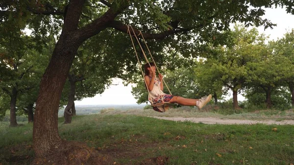 Vrij jong meisje swingend op een touw swingend op een eiken tak. gezond kind schommelend op schommel in park in de zon. Meisje houdt van vliegen op een schommel in het bos. concept van gelukkig gezin en jeugd. — Stockfoto