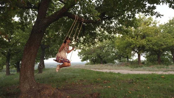 Gratis ung flicka svänger på ett rep svänger på en ek gren. friska barn svänger på swing i parken i solen. Flicka älskar att flyga på en gunga i skogen. begreppet lycklig familj och barndom. — Stockfoto