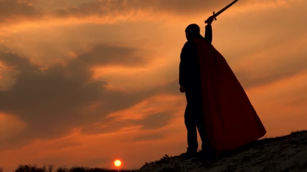 Воин в красном плаще стоит на горе и машет мечом в лучах заката. свободный рыцарь-мужчина. свободный человек играет супергероя. игра римского легиона в ярких лучах солнца против неба — стоковое видео
