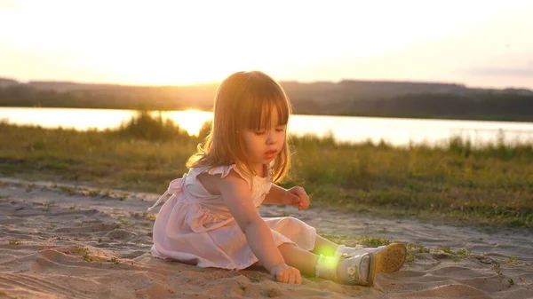 Die kleine Tochter sammelt bei Sonnenuntergang Sand auf. Das gesunde Baby spielt im Sand am Strand. glückliches Mädchen spielt im Park in der Sonne. Urlaubs- und Reisekonzept. Kind im Sommer auf der Wiese. — Stockfoto