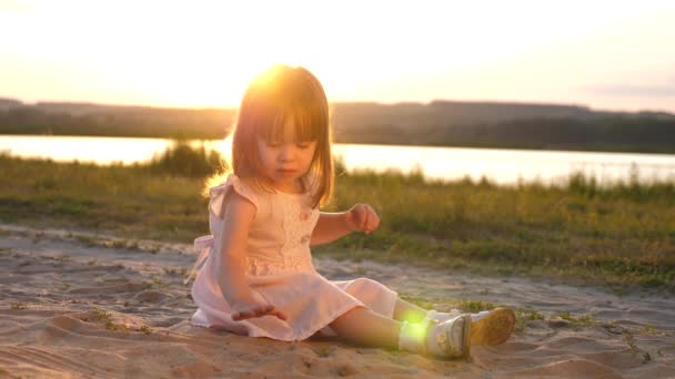 La figlioletta raccoglie sabbia al tramonto. bambino sano gioca nella sabbia sulla spiaggia. ragazza felice gioca nel parco al sole. Vacanza e concetto di viaggio. bambino in estate sul prato . — Video Stock