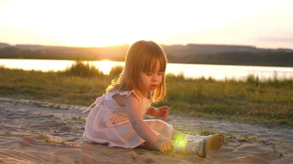 Die kleine Tochter sammelt bei Sonnenuntergang Sand auf. Das gesunde Baby spielt im Sand am Strand. glückliches Mädchen spielt im Park in der Sonne. Urlaubs- und Reisekonzept. Kind im Sommer auf der Wiese. — Stockfoto