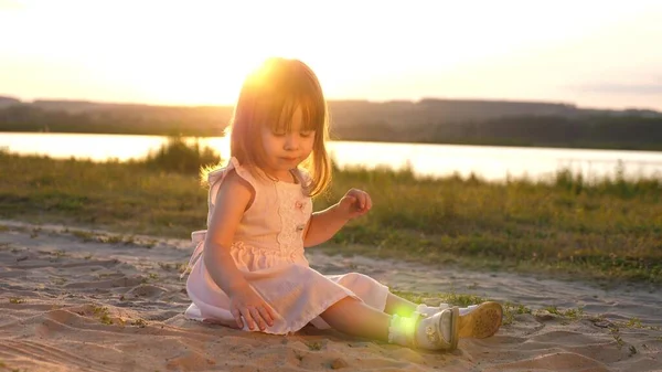 Die kleine Tochter sammelt bei Sonnenuntergang Sand auf. Das gesunde Baby spielt im Sand am Strand. glückliches Mädchen spielt im Park in der Sonne. Urlaubs- und Reisekonzept. Kind im Sommer auf der Wiese. — Stockfoto