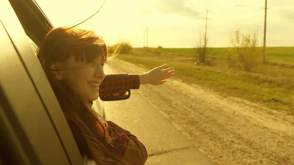Gratis kvinna reser med bil fånga vinden med handen från bilfönstret. Flicka med långt hår sitter i framsätet av bilen, sträcker ut armen genom fönstret och fånga bländning av nedgående sol — Stockfoto