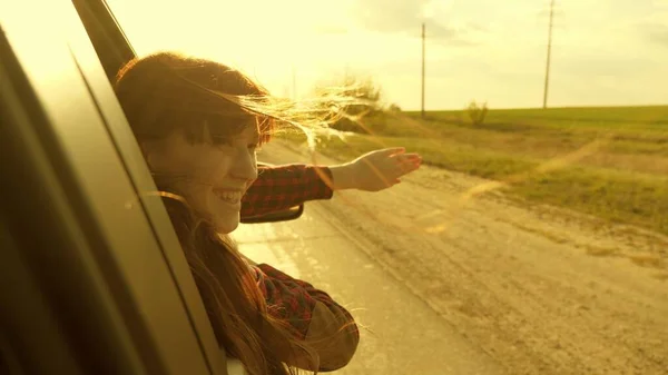 Gratis kvinna reser med bil fånga vinden med handen från bilfönstret. Flicka med långt hår sitter i framsätet av bilen, sträcker ut armen genom fönstret och fånga bländning av nedgående sol — Stockfoto