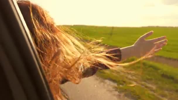 Dívka s dlouhými vlasy sedí na předním sedadle auta, natahuje ruku ven z okna a zachytává oslnivý pohled zapadající bezslunné ženy, která cestuje autem, chytá vítr rukou z okna auta. dobrodružství a — Stock video