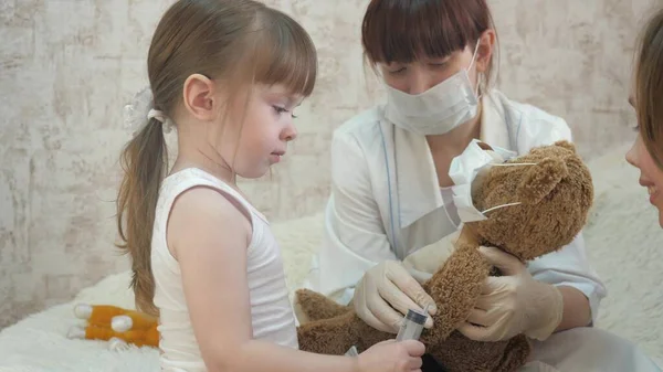 As crianças brincam com uma seringa médica com uma máscara protetora. jogo finge ser médico, enfermeiro, trata paciente com vacina. menina saudável injeta brinquedo urso. criança brinca no hospital . — Fotografia de Stock