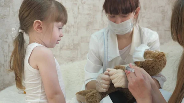 A gyermekek védőmaszkban lévő orvosi fecskendővel játszanak. játék úgy tesz, mintha orvos, nővér, kezeli beteg vakcinát. egészséges kislány játékmacit fecskendez be. gyermek játszik a kórházban. — Stock Fotó
