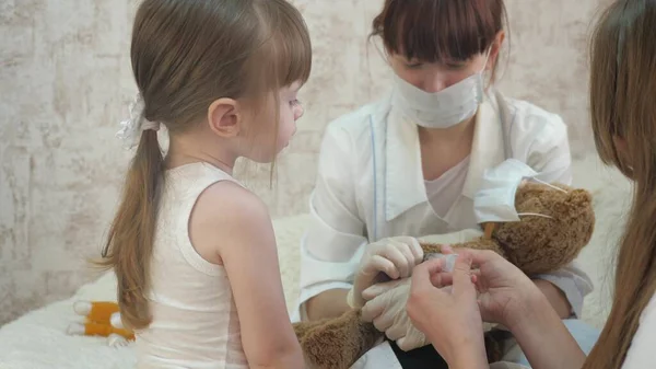子供たちは医療用注射器で保護マスクをして遊ぶ。ゲームは医者、看護師、ワクチンで患者を治療するふりをします。健康な女の子はおもちゃのクマを注入します。子供は病院で. — ストック写真