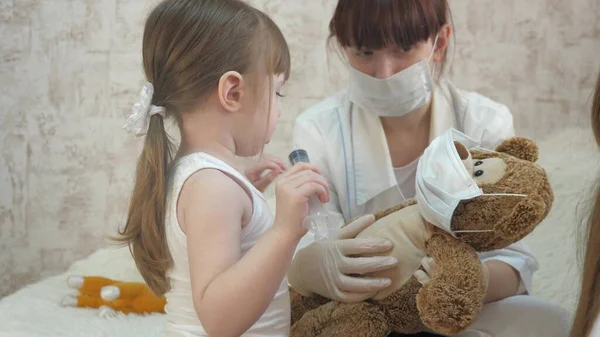 A gyermekek védőmaszkban lévő orvosi fecskendővel játszanak. játék úgy tesz, mintha orvos, nővér, kezeli beteg vakcinát. egészséges kislány játékmacit fecskendez be. gyermek játszik a kórházban. — Stock Fotó