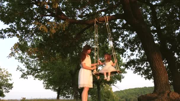 Mom shakes a healthy little daughter on swing under tree in sun. mother plays with child they are swinging on a rope on an oak branch in forest. girl laughs, rejoices. Free family having fun in park — Stock Video