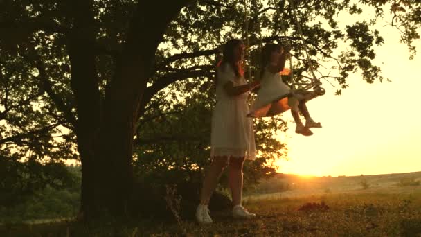 Мама качает здоровую маленькую дочь на качелях под солнцем. Мать играет с ребёнком, они висят на веревке на дубовой ветке в лесу. Свободная семья веселится в парке. счастливое детство, семья — стоковое видео