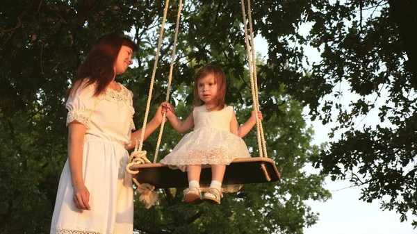 Een moeder is aan het spelen met een kind ze zwaaien aan een touw op een eik tak in het bos. gratis familie heeft plezier in Park. moeder rotsen gezond dochtertje op swing onder boom in de zon. — Stockfoto