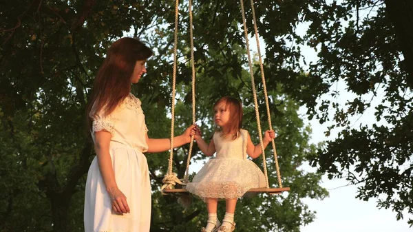 A mother is playing with child they are swinging on rope on an oak branch in forest. free family has fun in Park. mother rocks healthy little daughter on swing under tree in sun. — Stock Photo, Image