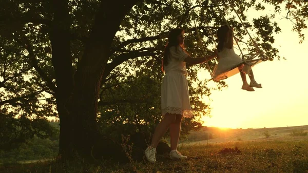 Mamma rockar frisk liten dotter på swing under träd i solen. en mamma leker med barnet de svingar på rep på en ekgren i skogen. gratis familj har kul i Park. lycklig barndom, familj — Stockfoto