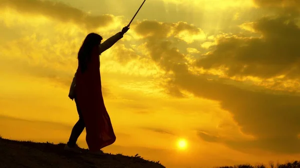 Super mulher em um manto vermelho acena uma espada sonha em lutar em uma montanha na luz do pôr do sol. Mulher livre a fazer de super-herói. menina joga lênin romano em raios brilhantes do sol contra o céu — Fotografia de Stock