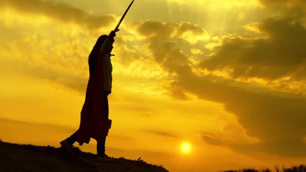 Super mujer en una capa roja ondea una espada sueña con luchar en una montaña en la luz del atardecer. mujer libre jugando a ser un superhéroe. chica juega lenin romano en los rayos brillantes del sol contra el cielo — Foto de Stock