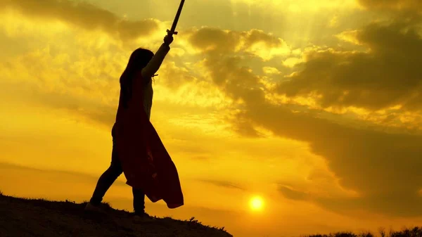 Super kvinna i en röd mantel vinkar ett svärd drömmar om att slåss på ett berg i solnedgången ljus. gratis kvinna spelar en superhjälte. flicka spelar romersk Lenin i ljusa solstrålar mot himlen — Stockfoto