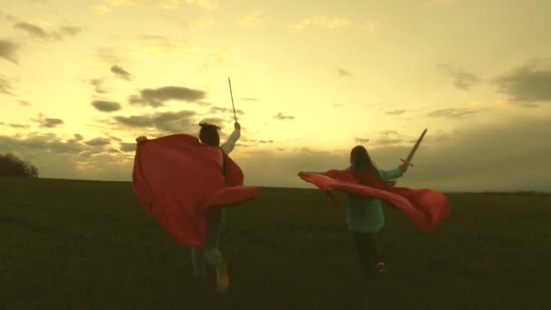 穿着红色斗篷的女孩手拿剑穿过田野，玩中世纪骑士游戏。孩子们用玩具剑战斗。孩子们玩骑士游戏。快乐童年的概念。健康的年轻女孩玩超级棒 — 图库视频影像