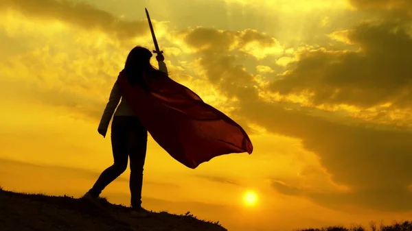 Super vrouw in een rode mantel zwaait een zwaard droomt van vechten op een berg in de zonsondergang licht. vrije vrouw die een superheld speelt. meisje speelt roman lenin in heldere stralen van de zon tegen de hemel — Stockfoto