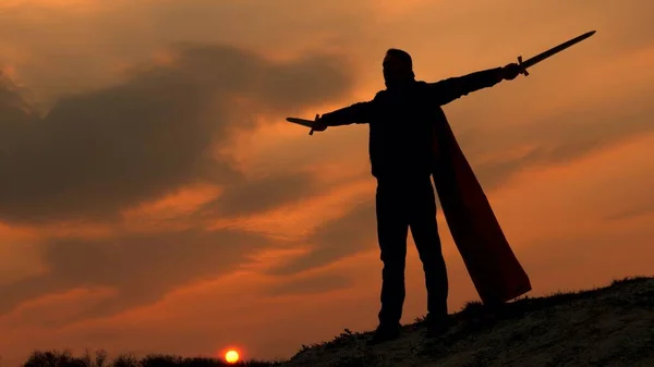 Free man plays superhero. game of Roman legion. free male knight prays with swords outstretched hands. Superman with a sword in his hand and in a red cloak stands on mountain in sunset light. — Stock Photo, Image