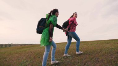 Turist kızlar tepeye tırmanan çok renkli yağmurluklarla sırt çantasıyla seyahat ederler. Özgür kadın gezginler el ele dağın zirvesine çıkar. Ağır çekim. Takım çalışması gezginleri. macera konsepti.