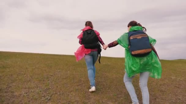 健康的女孩带着背包旅行，穿着五颜六色的雨衣爬山。自由女旅行家手牵着手爬上山顶。慢动作。团队旅行。冒险概念. — 图库视频影像