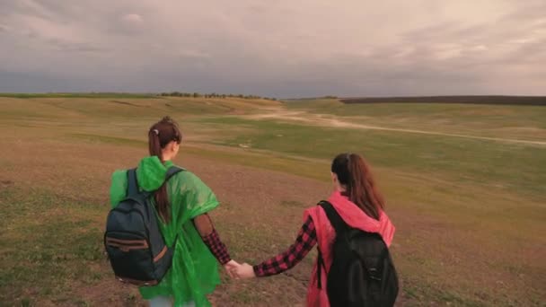 Avontuur en reis concept. vrije vrouwen reizigers dalen af van heuvel naar een prachtige vallei hand in hand. gezonde meisjes toeristen reizen met rugzakken in veelkleurige regenjassen. teamwork reizigers. — Stockvideo