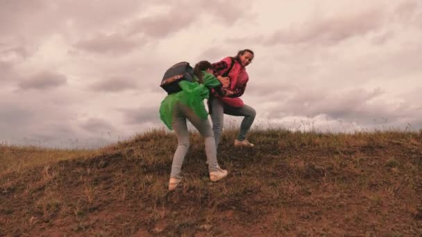 Zdrowe dziewczyny turystyczne podróżują w kolorowych płaszczach przeciwdeszczowych, wspinają się na wzgórze trzymając się za ręce, pomagają sobie nawzajem. wolne kobiety z plecakami podróżnicy iść na szczyt góry, radować się zwycięstwem i skakać. praca zespołowa — Wideo stockowe