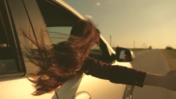 Ελεύθερη γυναίκα ταξιδεύει με το αυτοκίνητο πιάνει τον άνεμο με το χέρι της από το παράθυρο του αυτοκινήτου. Κορίτσι με μακριά μαλλιά κάθεται στο μπροστινό κάθισμα του αυτοκινήτου, τεντώνοντας το χέρι της έξω από το παράθυρο και να πιάσει λάμψη από τη δύση του ήλιου — Αρχείο Βίντεο