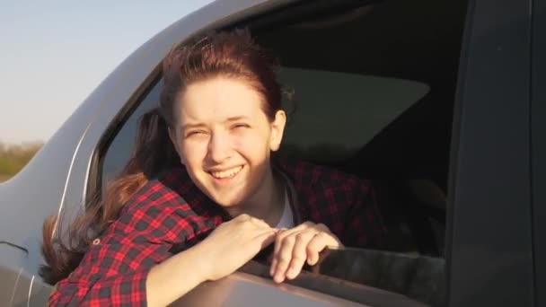 Una mujer libre viaja en coche, se regocija en el viento desde la ventana del coche. Una chica con el pelo largo se sienta en el asiento trasero de un coche, mira por una ventana y sonríe al sol. concepto de aventura de coche — Vídeos de Stock