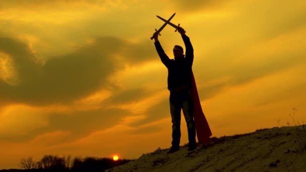Chevalier mâle libre a croisé ses épées dans le ciel au-dessus de sa tête. Superman avec une épée à la main et dans un manteau rouge se tient sur la montagne dans la lumière du coucher du soleil. homme libre joue un super-héros. jeu de légion romaine . — Video