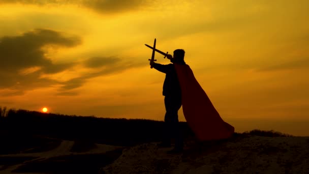 Ελεύθερος ιππότης διέσχισε τα σπαθιά του στον ουρανό πάνω από το κεφάλι του. Σούπερμαν με ένα σπαθί στο χέρι του και σε ένα κόκκινο μανδύα στέκεται στο βουνό στο φως του ηλιοβασιλέματος. Ο ελεύθερος άνθρωπος παίζει τον υπερήρωα. παιχνίδι της ρωμαϊκής λεγεώνας. — Αρχείο Βίντεο