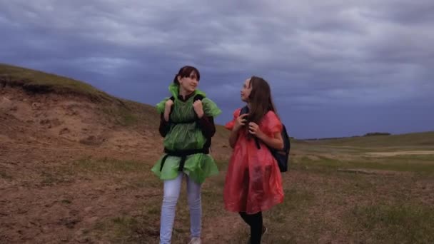 Zdrowe dziewczyny turystyczne podróżują z plecakami w kolorowych płaszczach przeciwdeszczowych, zbliża się burza. Wolne kobiety idą do wąwozu, trzymając się za ręce. podróżnych do pracy zespołowej. Koncepcja przygody i podróży. — Wideo stockowe