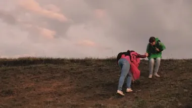 Özgür kadın gezginler tepenin tepesinden el ele tutuşarak inerler. Takım çalışması gezginleri. Macera ve seyahat kavramı. Sağlıklı turist kızları renkli yağmurluklarla sırt çantalarıyla seyahat ederler..