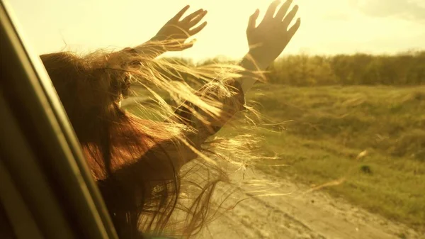 Donna libera viaggia in auto cattura il vento con la mano dal finestrino dell'auto. Ragazza con i capelli lunghi è seduto sul sedile anteriore della macchina, allungando il braccio fuori finestra e la cattura bagliore del sole al tramonto — Foto Stock