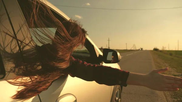 Femme libre voyage en voiture attrape le vent avec sa main par la fenêtre de la voiture. Fille aux cheveux longs est assis dans le siège avant de la voiture, étirant son bras par la fenêtre et attrapant l'éblouissement du soleil couchant — Photo