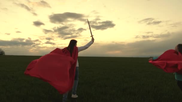 Gratis unga flickor spela superhjältar. Flickor i röda mantlar springer med svärd i hand över fältet och leker medeltida riddare. Barn slåss med leksakssvärd. Friska barn leker riddare. barndomsbegrepp. — Stockvideo