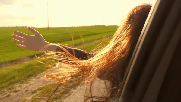 Donna libera viaggia in auto cattura il vento con la mano dal finestrino dell'auto. Ragazza con i capelli lunghi è seduto sul sedile anteriore della macchina, allungando il braccio fuori finestra e la cattura bagliore del sole al tramonto — Foto Stock