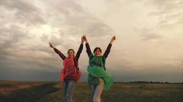 strong and healthy girls travelers travel in colorful raincoats, climb mountain, hold hands, help each other. Free women tourists with backpacks on rise to top of hill, rejoice in victory and jump