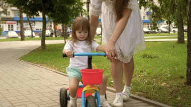 Sağlıklı çocuk bisiklet sürmeyi öğrenir. Ebeveynler ve küçük kızları parkta yürürler. Mutlu anne küçük kızına bisiklet sürmeyi öğretiyor. Anne bebeğiyle dışarıda oynuyor. Mutlu aile ve çocukluk kavramı