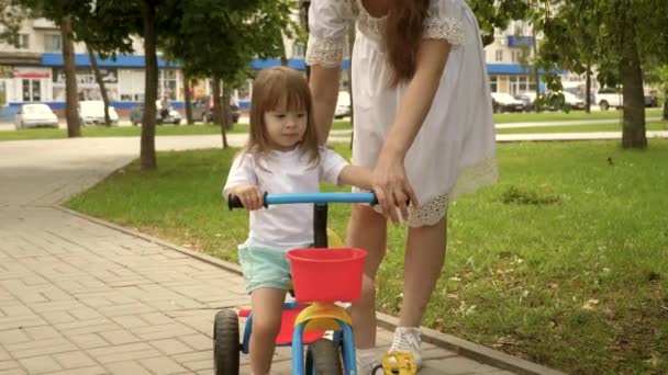 Sağlıklı çocuk bisiklet sürmeyi öğrenir. Ebeveynler ve küçük kızları parkta yürürler. Mutlu anne küçük kızına bisiklet sürmeyi öğretiyor. Anne bebeğiyle dışarıda oynuyor. Mutlu aile ve çocukluk kavramı — Stok video