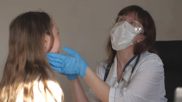 ゴーグルと医療用手袋を装着した医療用マスクの医師は、子供の喉と目を検査します。白いコートと聴診器を着た女が少女の健康をチェックしてる。病院病棟の小児科医と. — ストック写真