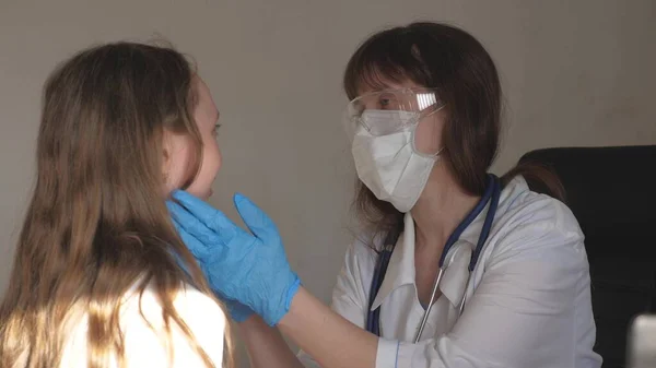 En läkare i medicinsk mask med skyddsglasögon och medicinska handskar undersöker halsen och ögonen på barn. kvinna i vit rock och stetoskop kontrollerar hälsan hos liten flicka. barn på sjukhus avdelning med läkare. — Stockfoto