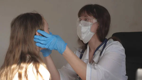 ゴーグルと医療用手袋を装着した医療用マスクの医師は、子供の喉と目を検査します。白いコートと聴診器を着た女が少女の健康をチェックしてる。病院病棟の小児科医と. — ストック写真