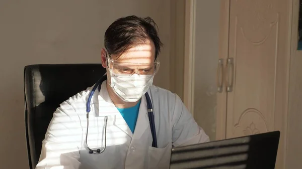 Doctor en consultorio en bata blanca y gafas de seguridad trabajando en laptop, consulta online y conferencia. El médico se comunica con un cliente mediante chat virtual. Telemedicina, atención médica remota — Foto de Stock