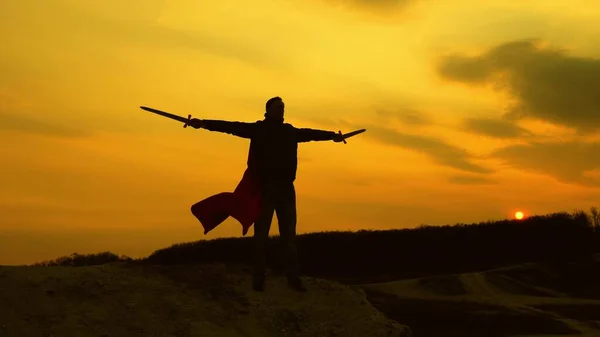 Un guerrero con una espada en la mano y con una capa roja está parado en la montaña en la luz del atardecer. caballero masculino libre reza con espadas extendidas manos. hombre libre juega superhéroe. juego de la legión romana . — Foto de Stock